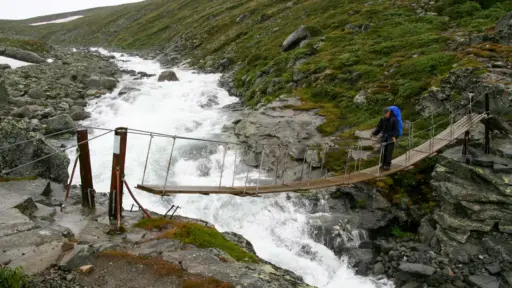 Schmale Hängebrücke im Jotunheimen-Nationalpark – Wanderer mit Trekkingrucksack bei Regen Schmale Hängebrücke im Jotunheimen-Nationalpark in Norwegen, ein Wanderer mit Trekkingrucksack und Regenhülle überquert sie bei Regen.