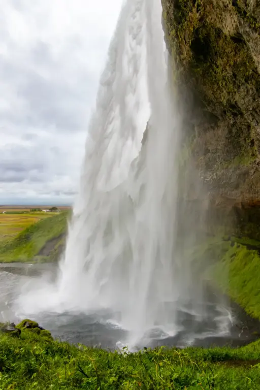 Seljalandsfoss-wasserfall-pfad-rueckseite Seitenansicht des Seljalandsfoss-Wasserfalls in Island, fotografiert vom begehbaren Pfad direkt hinter den herabstürzenden Wassermassen vor einer grünen Landschaft unter bewölktem Himmel.