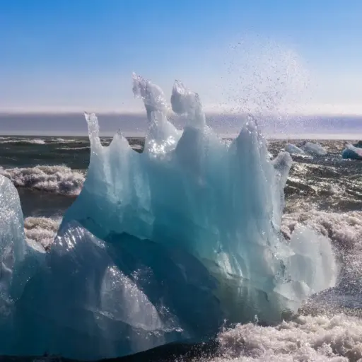 Eisskulptur in der Brandung am Diamond Beach Ein gläserner, bizarr geformter Eisblock am schwarzen Sandstrand Diamond Beach, der von einer Meereswelle umspült wird, während Gischt in der Sonne glitzert.