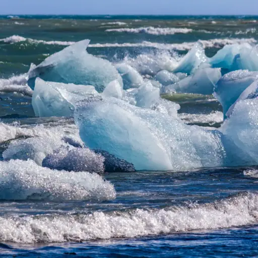 Eis-Diamanten am Diamond Beach Kristallklare, blau schimmernde Eisblöcke in der Brandung an einem schwarzen Sandstrand unter blauem Himmel.