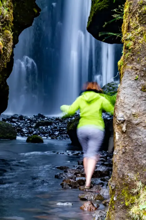 Der Weg in die Gljúfrabúi-Schlucht Rückansicht einer Person in einer neongrünen Jacke, die sich durch Bewegungsunschärfe unscharf abhebt, während sie über Steine im Bachbett in die enge Felsenschlucht des Gljúfrabúi Wasserfalls in Island wandert. Im Hintergrund stürzt das Wasser als weicher Schleier herab.