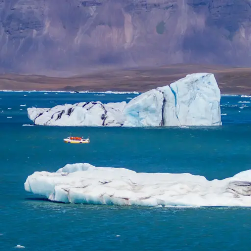 Bootstour in der Jökulsárlón-Lagune Ein gelbes Amphibienfahrzeug mit Touristen fährt in einer blauen Lagune zwischen riesigen, weiß-blauen Eisbergen vor einer bergigen Kulisse.
