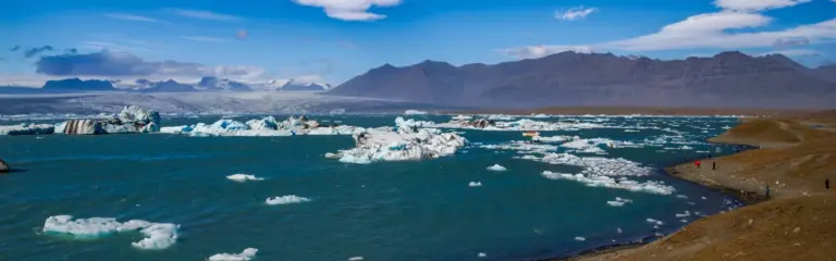 Jökulsárlón Gletscherlagune, Island Türkisfarbene Gletscherlagune in Island mit schwimmenden Eisbergen, klein wirkenden Ausflugsbooten, und Bergen im Hintergrund unter blauem Himmel.