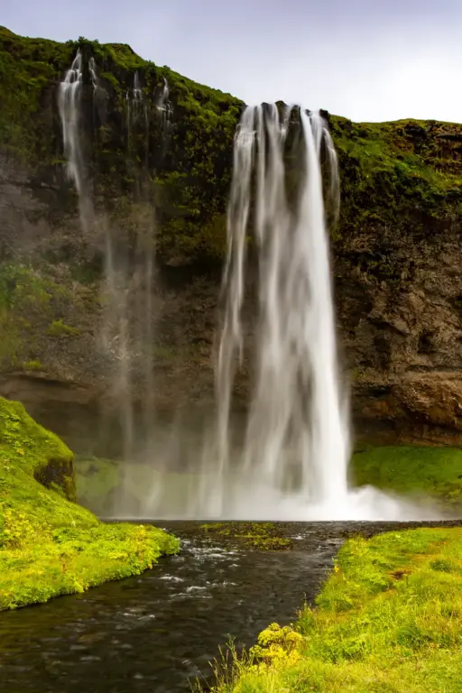 Der Seljalandsfoss in seiner vollen Pracht Frontale Totalaufnahme des Seljalandsfoss-Wasserfalls in Island, bei der das Wasser über eine moosbewachsene Klippe in einen Fluss stürzt, der im Vordergrund durch grüne Wiesen fließt.