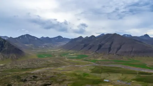 Die Berge der Westfjorde bei Þingeyri Ein weites, grünes Tal in den isländischen Westfjorden, umgeben von steilen, dunklen Bergketten unter einem bewölkten Himmel.