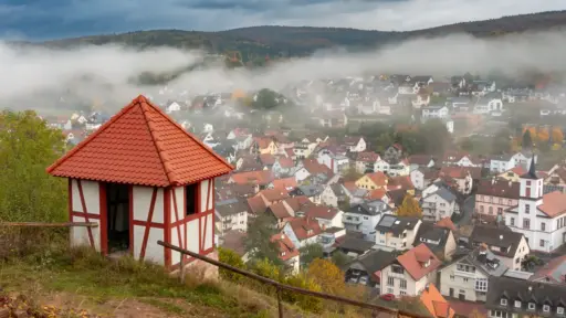 Partenstein von oben links ein wiederaufgebautes Wachhäuschen der Burgruine Partenstein mit rotem Fachwerk, dahinter im nebelverhangendem Tal der Ort Partenstein