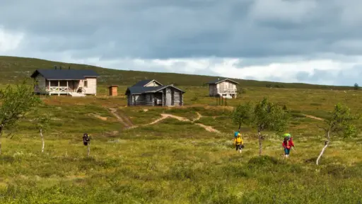 kuivin-autiotupa-wildnishuette-trekking-finnland Drei Wanderer mit Rucksäcken laufen durch die weite Fjäll-Landschaft Lapplands bei einer Wildnishütte.