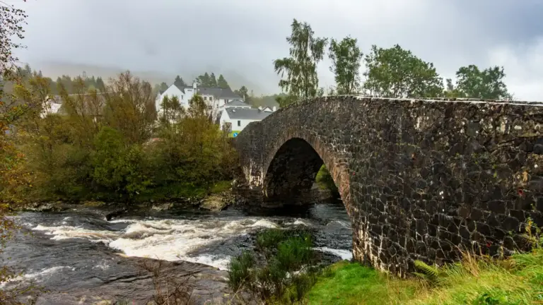 Die historische Brücke in Bridge of Orchy, West Highland Way Eine alte Bogenbrücke aus dunklem Stein führt über einen schäumenden Fluss in den schottischen Highlands, im Hintergrund steht das weiße Bridge of Orchy Hotel unter wolkenverhangenem Himmel.