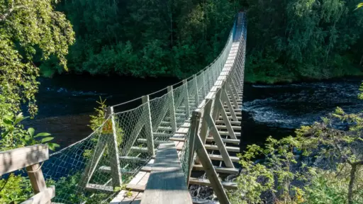 Hängebrücke über Stromschnellen des Oulankajoki lange Hängebrücke über einen Fluss mit Stromschnellen. im Hintergrund dichter Wald im Schatten.