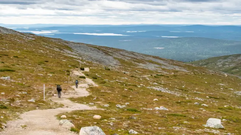 Wanderer auf einem breiten absteigenden Wanderweg im weiten Fjell. Im Hintergrund bewölkter Himmel über der weiten Landschaft mit zahlreichen Seen.