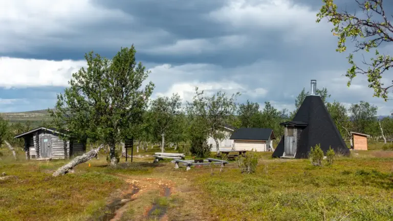 Hüttenstation Njávgoaivi im Paistunturi-Wildnisgebiet Wildnishütte, Kota, Holzlager, Trockentoillete und Feuerstellen auf einer Wiese mit lichten Birken unter bewölktem Himmel,