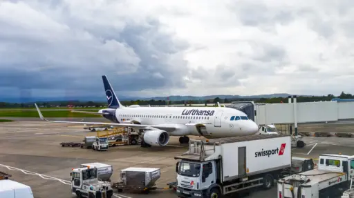 A320 von Lufthansa am Flughafen Glasgow Flugzeug bei bewölktem Himmel auf dem Rollfeld mit Passagiertunnel und Gepäcktransport-Einheiten.