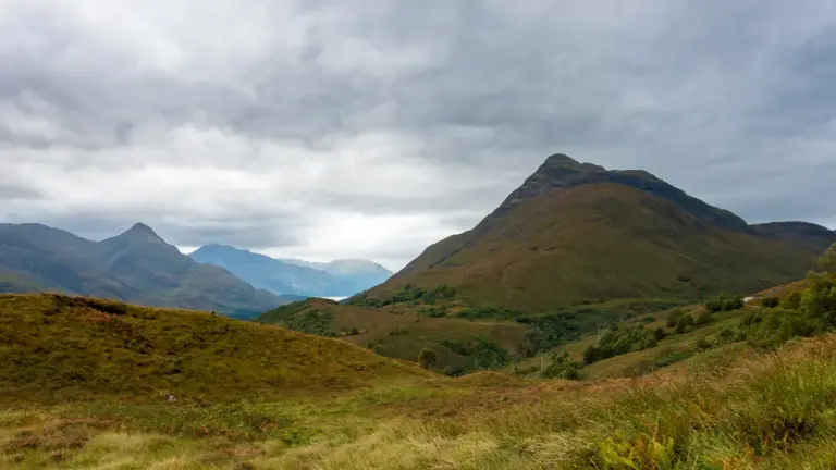 Beinn na Caillich (762 m) bei wolkenverhangenem Himmel Blick von einem erhöhten, grasbewachsenen Berghang über ein tiefes Tal nach Westen. Auf der rechten Seite ragt ein mächtiger, steiler Bergrücken auf. In der Ferne schimmert das Wasser des Loch Leven zwischen den umliegenden Berghängen unter einem wolkenverhangenen, grauen Himmel.