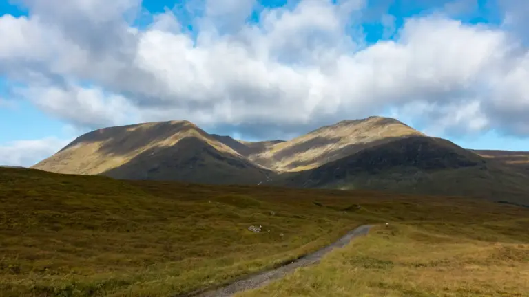 Bergpanorama des Black Mount Massivs Ein schottriger Wanderweg schlängelt sich durch eine weite, hügelige Graslandschaft hin zu zwei mächtigen, kahlen Berggipfeln im Hintergrund, deren Hänge von einem Wechselspiel aus intensivem Sonnenlicht und tiefen Wolkenschatten geprägt sind.