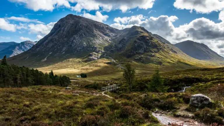 Buachaille Etive Mòr und Lagangarbh Hut Im Vordergrund führt ein schmaler Pfad zu einer kleinen Holzbrücke über einen Bach in einer hügeligen Heide- und Moorlandschaft. Dahinter ragt die gewaltige, steile Felswand eines Berges auf, an dessen Fuß ein einzelnes, kleines weißes Haus inmitten grüner Wiesen steht, während Wolkenschatten über die Hänge ziehen.