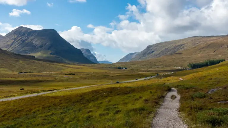 Buachaille Etive Mòr Massiv am Eingang zu Glen Coe Ein heller, schottriger Wanderweg führt durch eine weite, grüne Hochebene, parallel zu einer asphaltierten Straße mit vereinzelten Autos. Links ragt ein markanter, steiler Berggipfel auf, während im Hintergrund ein weites Tal unter blauem Himmel mit weißen Quellwolken liegt.