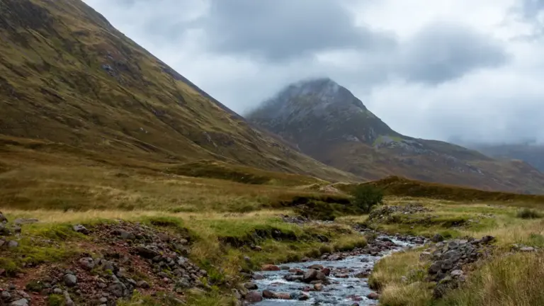 Bergbach Allt Coire na Sleubhauch im Lairigmor-Tal Ein felsiger, klarer Bergbach schlängelt sich im Vordergrund durch eine grüne, raue Graslandschaft. Im Hintergrund ragt ein steiler, herbstlich brauner Berghang empor, dessen spitzer Gipfel mystisch von tief hängenden, grauen Regenwolken eingehüllt wird.