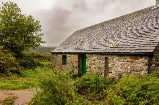 Die Doune Byre Bothy am Loch Lomond, West Highland Way Die urige Doune Byre Bothy aus rauem Naturstein mit einem Schieferdach und einer grünen Holztür steht inmitten von grünen Büschen direkt am Wanderpfad des West Highland Way.