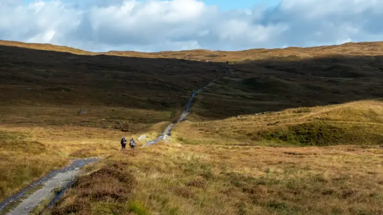 Rannoch Moor an der Old Military Road Zwei Wanderer mit großen Rucksäcken folgen einem schmalen, steinigen Pfad, der sich durch die hügelige, herbstlich braune Gras- und Heidekarglandschaft zieht, während die Wolken am Himmel ein kontrastreiches Muster aus Licht und Schatten auf die Hügel werfen.