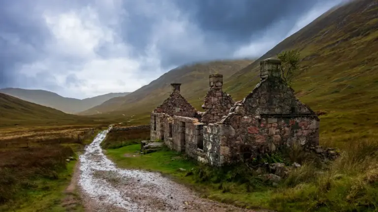 Ruine von Tigh-na-sleubhaich im Lairigmor-Tal am WHW Die steinernen Überreste des verlassenen Hauses Tigh-na-sleubhaich stehen an einer nassen Militärstraße in einem einsamen Tal der schottischen Highlands unter dunklen Regenwolken.
