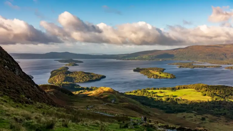 Panoramablick vom Conic Hill auf den Loch Lomond Blick von den Berghängen des Conic Hill auf den blauen Loch Lomond mit seinen bewaldeten Inseln unter einer dramatischen Wolkendecke im warmen Abendlicht.