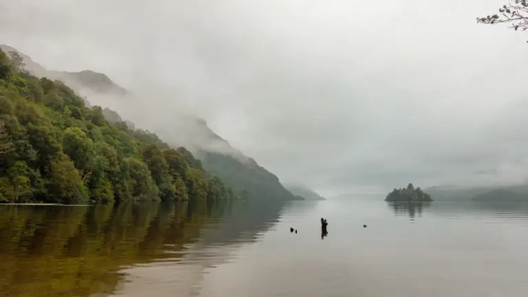 Nebelstimmung über dem Loch Lomond, West Highland Way Ein spiegelglatter, schottischer See im dichten Nebel, links ragt ein bewaldeter Berghang ins Wasser und rechts spiegelt sich eine kleine, grüne Insel im See.